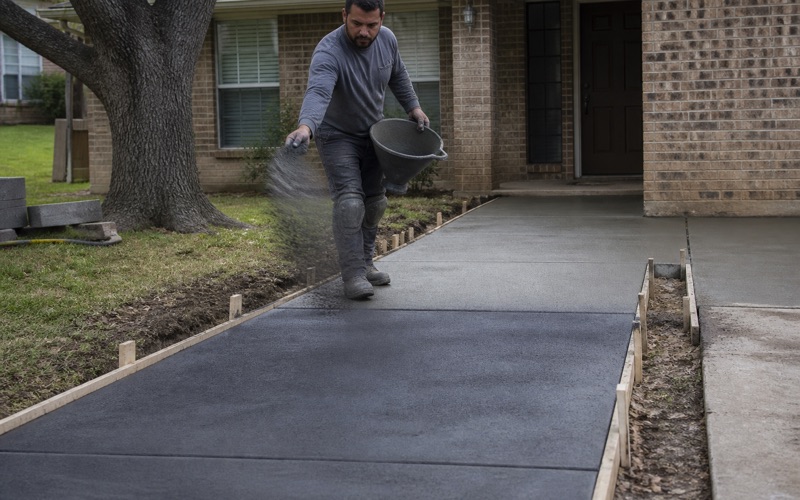 Charcoal color hardener being broadcast across a wet front entry walk in Killeen, the worked-in half meeting the bare gray half.